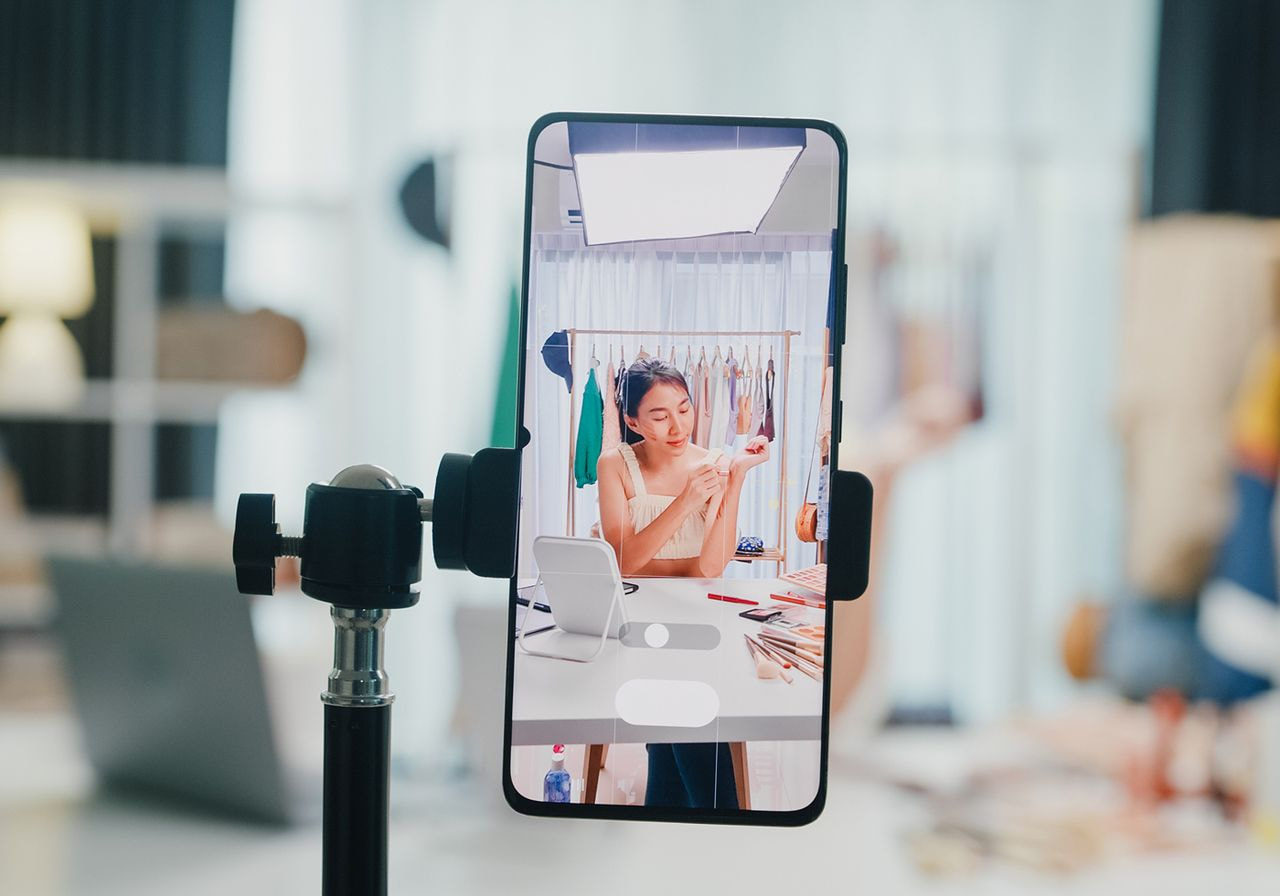 A smartphone on a tripod films a woman demonstrating makeup products at a table in front of a clothing rack and studio light.